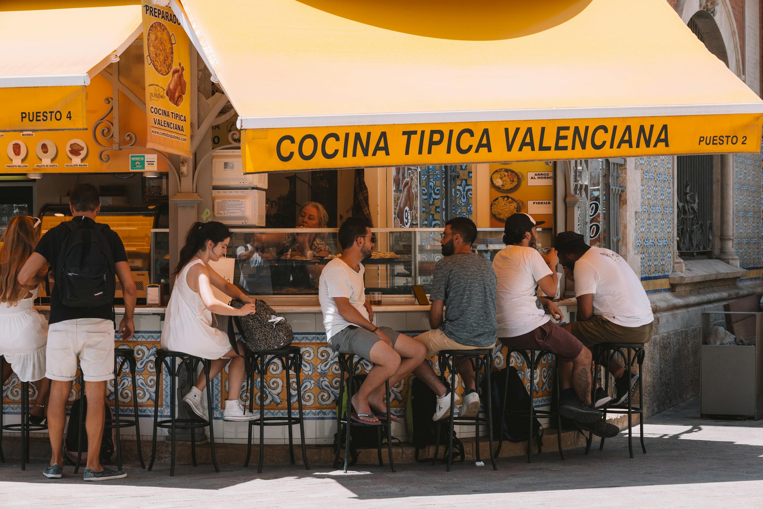 People enjoying food at a Valencian street stall under a bright yellow awning.